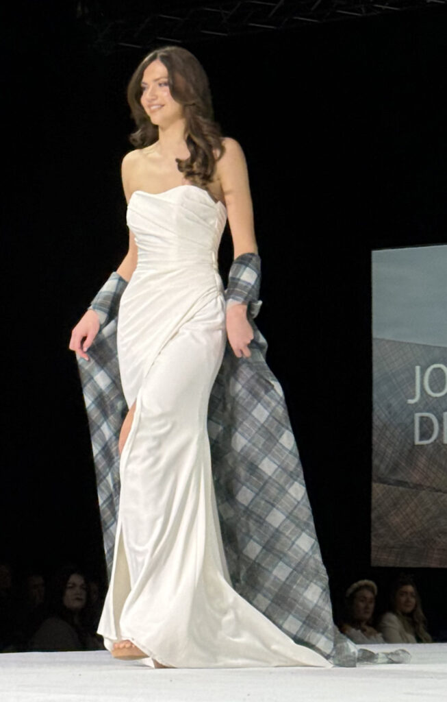 model in ivory wedding dress walking on catwalk with tartan organza cape flowing behind