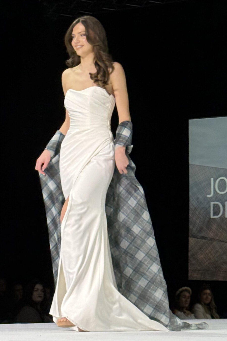 model in ivory wedding dress walking on catwalk with tartan organza cape flowing behind
