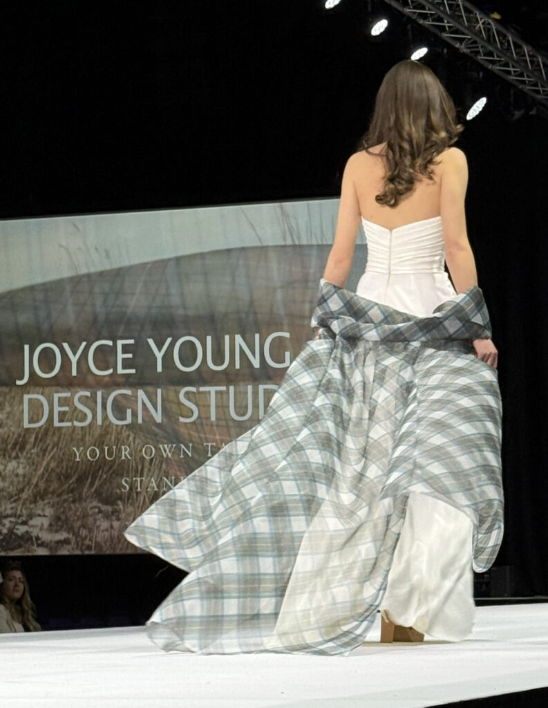 model in ivory wedding dress walking on catwalk with tartan organza cape flowing behind